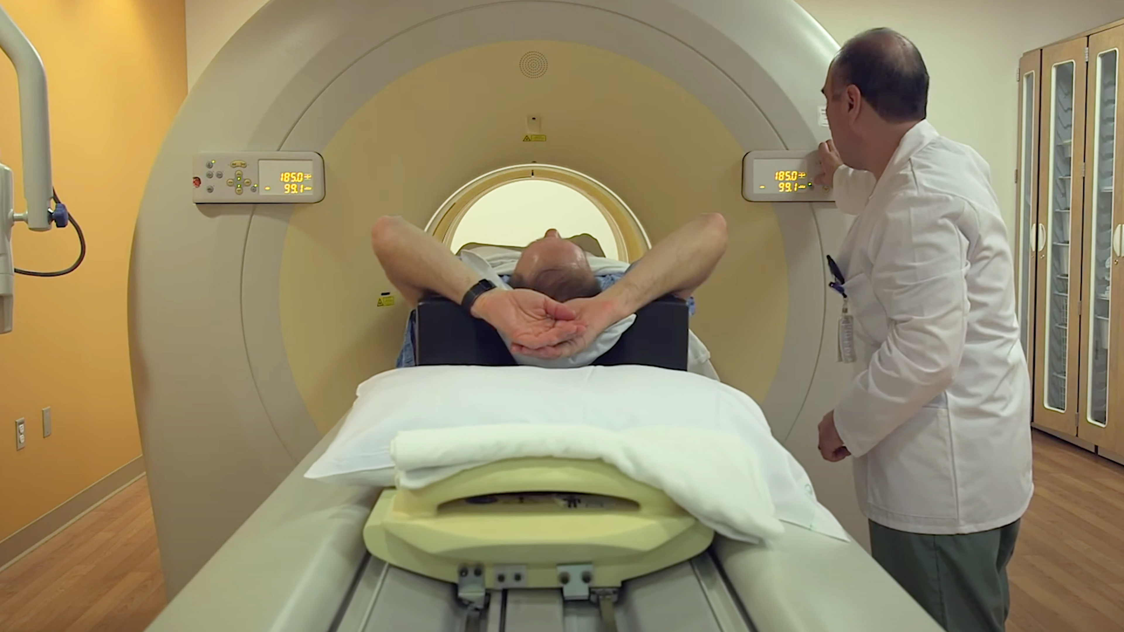 A patient lies comfortably on a CT scanner table with arms raised while a technologist assists nearby.